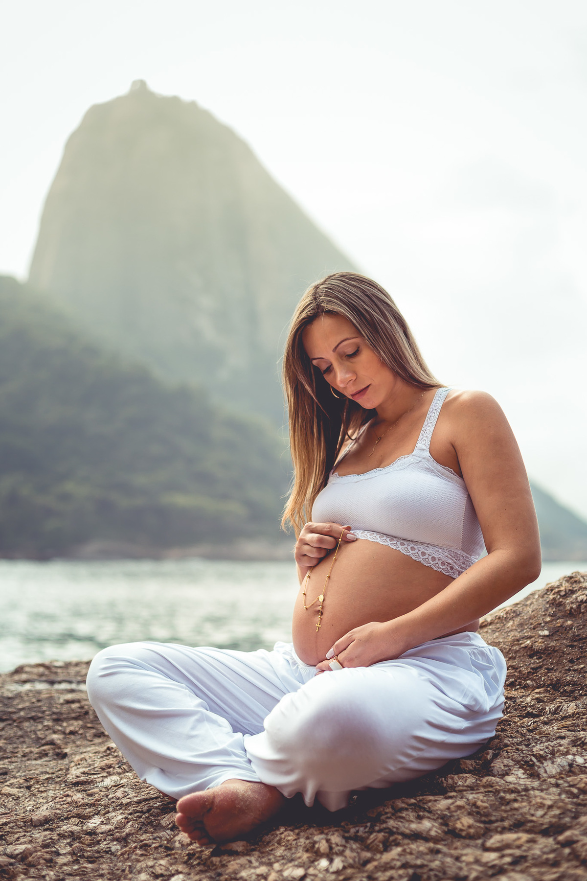 foto de gestante olhando para a barriga sentada numa pedra segurando um terço sobre a barriga e com pão de açucar ao fundo