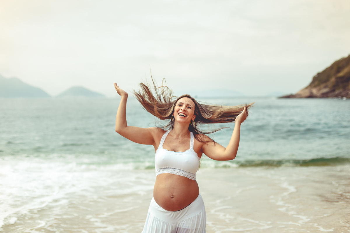 foto de grávida gargalhando em frente ao mar mexendo nos cabelos na praia vermelha