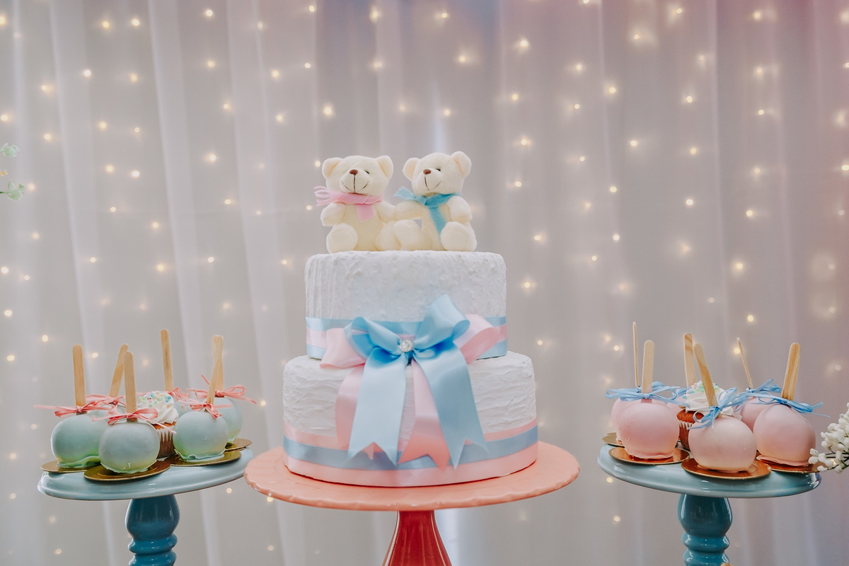 bolo branco com fitas de cetim rosa e azul com dois ursinhos no topo de bolo, ao fundo pontos de luz, ao lado do bolo maças nas cores azul e rosa