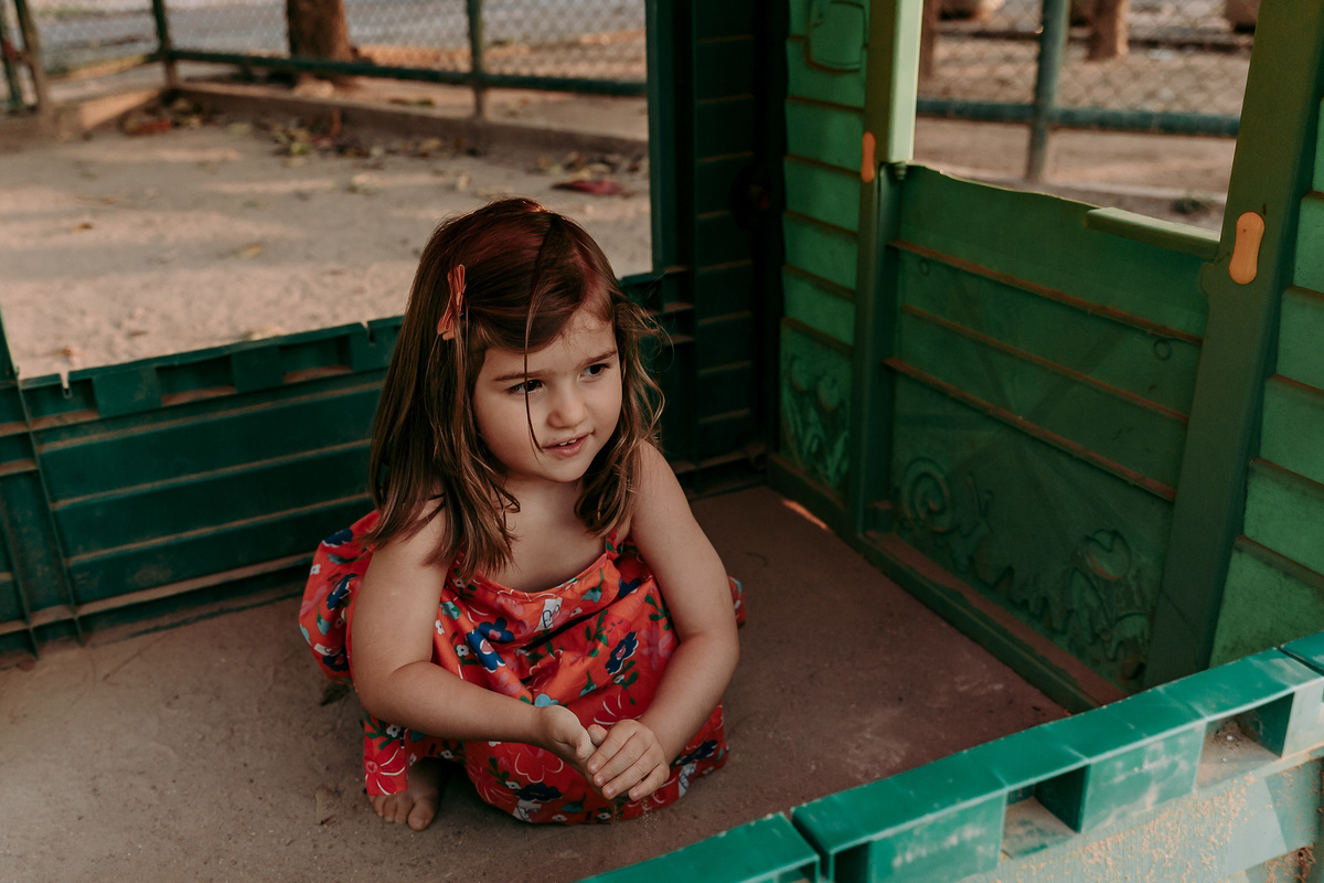 criança de vestido vermelho agachada em casinha de plástico em pracinha da urca durante ensaio família no rj