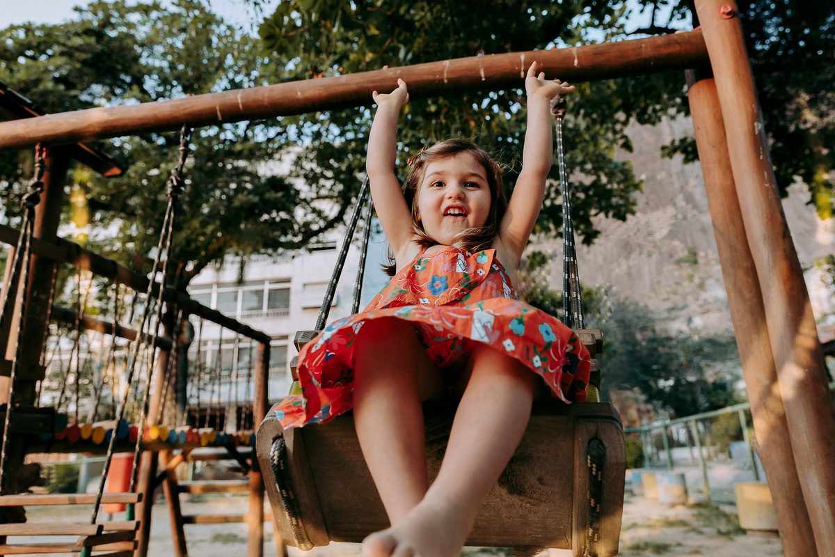 foto de criança de vestido vermelho com os braços levantados para o alto brincando alegremente em balanço de parquinho da Urca no rj