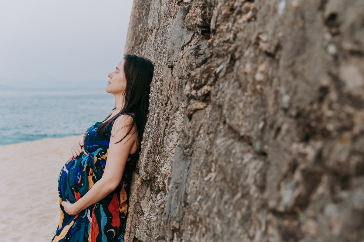 foto de gestante de vestido azul relaxada encostada em uma parede de pedra na praia vermelha no rj com as maos na barriga