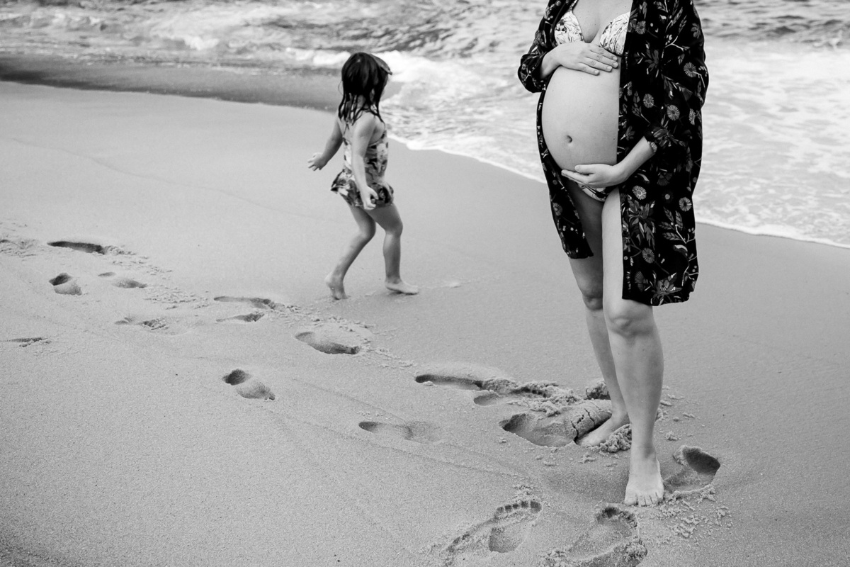 foto em preto e branco de barriga de gestante e sua filha correndo em direção ao mar da praia vermelha no rj