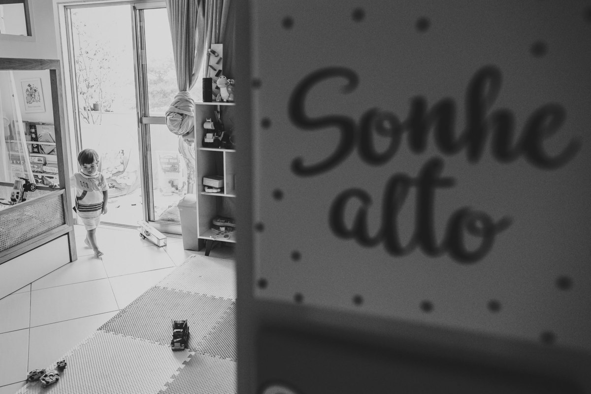 foto em preto e branco de criança em seu quarto e em primeiro plano a placa sonhe alto