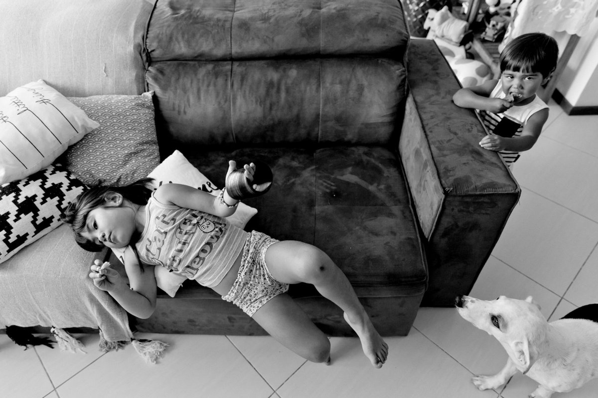 foto documental de duas crianças, uma esparramada no sofá e a outra em pé ao lado de um cachorro branco