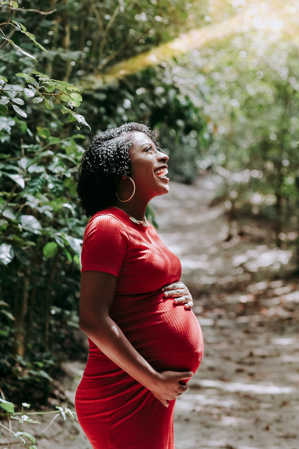 gestante de vestido vermelho e posicionada de lado para a camera sorri olhando para o ceu em seu ensaio gestante no parque chico mendes rj