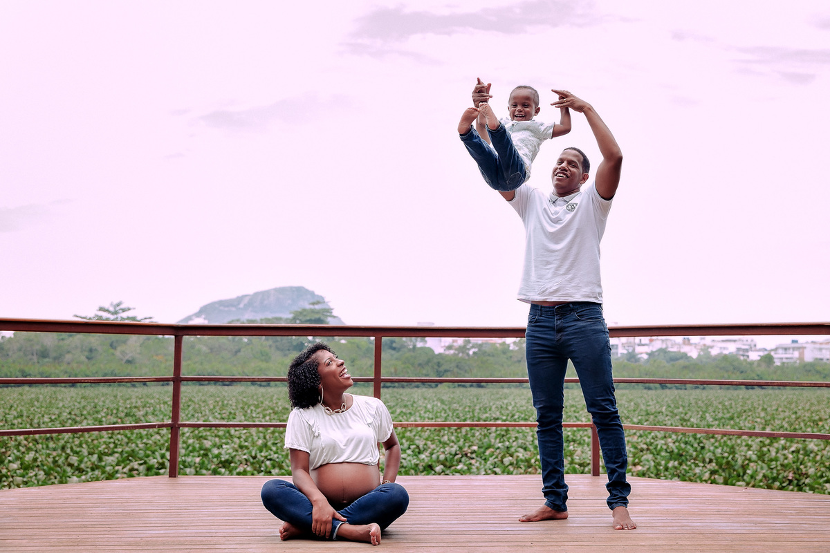 mae gestante sentada de pernas cruzadas no chão de deck do parque chico mende rj olhando sorrindo para o seu marido enquanto pai está ao lado brincando com seu filho de 2 anos jogando ele para o alto