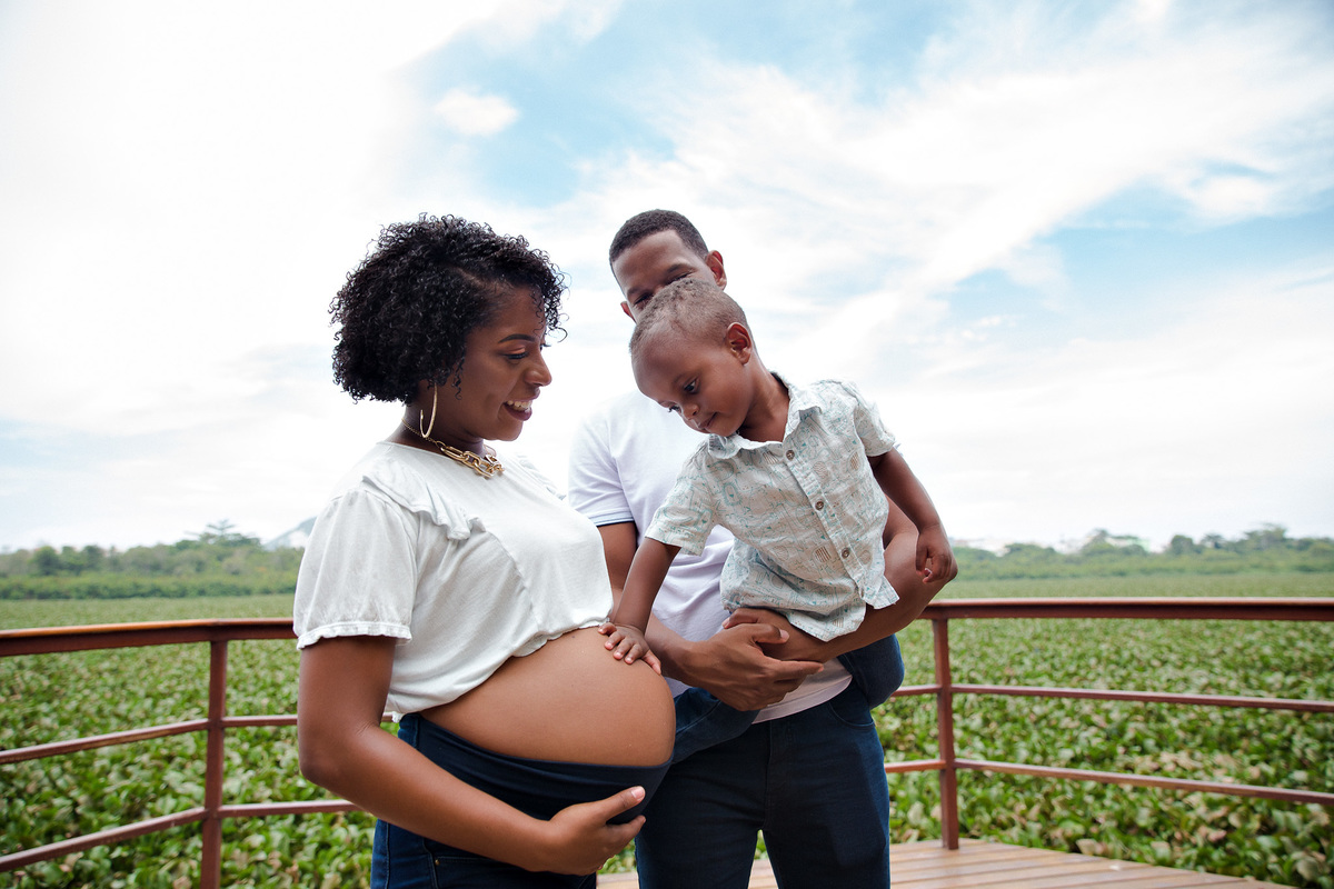 filho toca a barriga de sua mae que está gestante enquanto seu pai o segura no colo