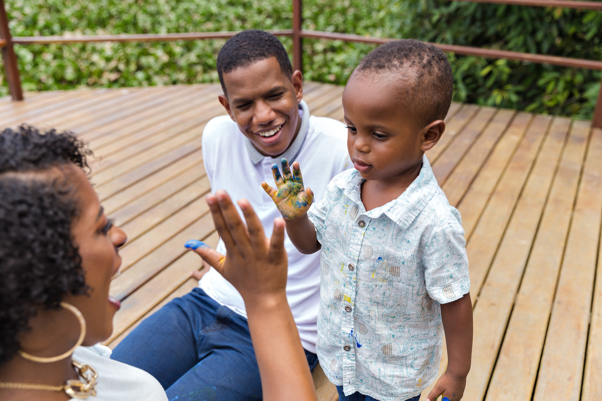 criança de 2 anos com a mao suja de tinta guache brinca de bater na mão de sua mae enquanto são observados pelo pai