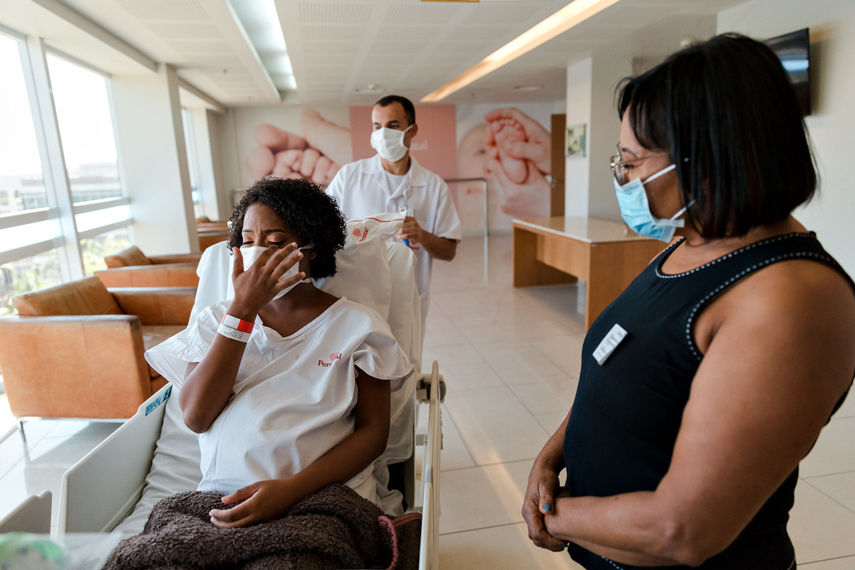 gestante deitada na maca chora emocionada minutos antes de seguir para centro cirurgico na perinatal da barra enquanto é observada por sua mae e maqueiro todos de máscara por conta da covid-19