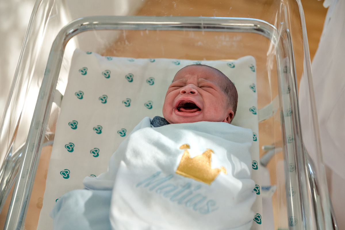 bebê  chorando embrulhado em manta na cor azul claro e bordada com uma cora de rei e seu nome matias, em berçario da perinatal da barra da tijuca rj