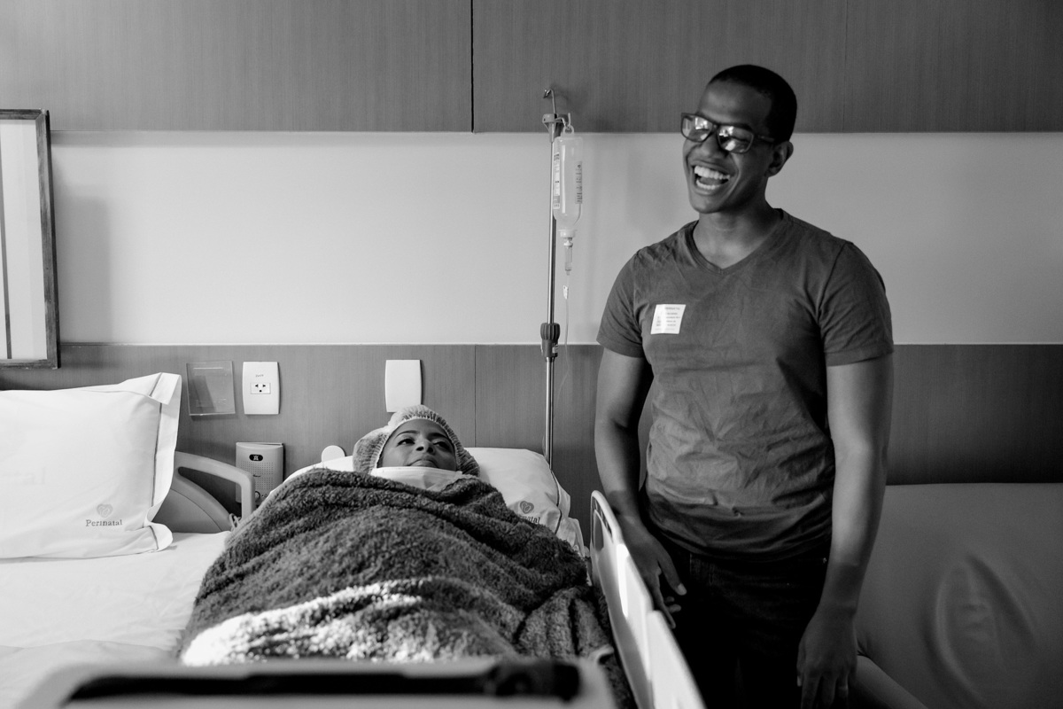 pai sorri ao lado de maca de hospital com sua esposa que acabou de dar a luz a seu filho