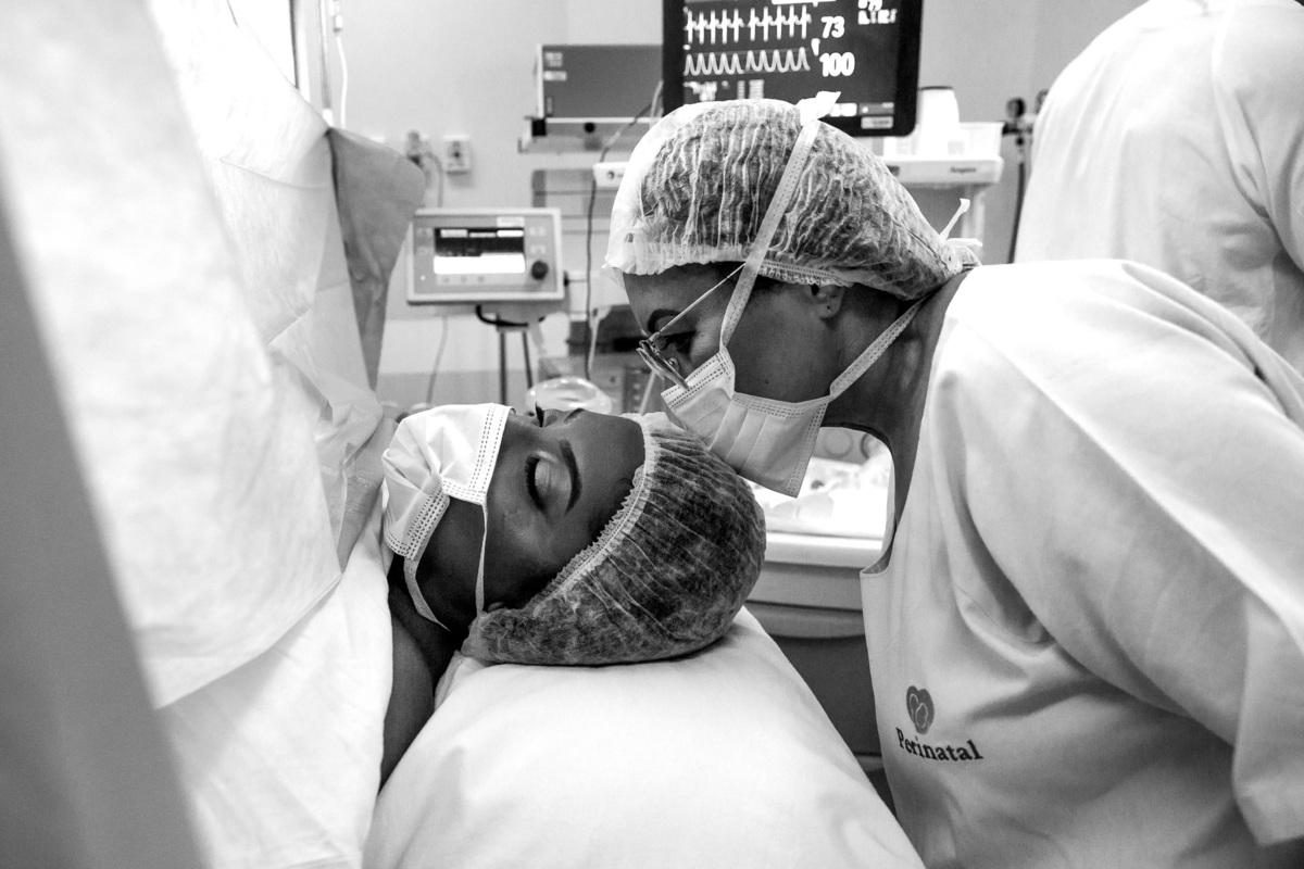 mae beija a testa da filha durante a cesariana, gestante de olhos fechados, ambas de mascara por conta da pandemia da covid-19