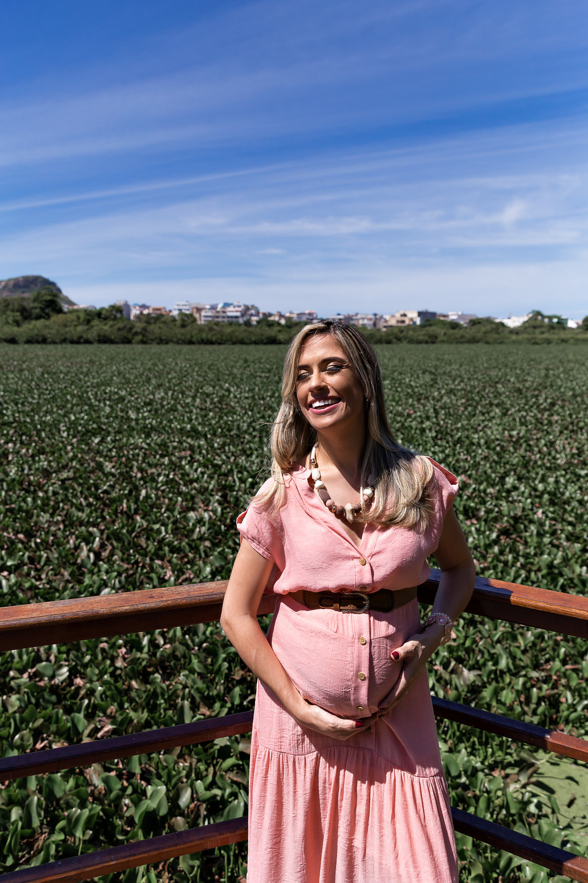 foto na vertical de mulher gestante de vestido rosa com um mar de gigoias ao fundo