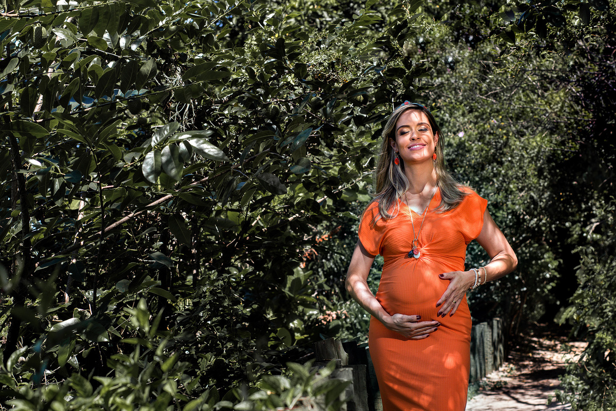 foto de gestante de vestido laranja em ambiente repleto de verde, com face voltada para o sol e discreto sorriso e com as duas maos acariando a sua barriga