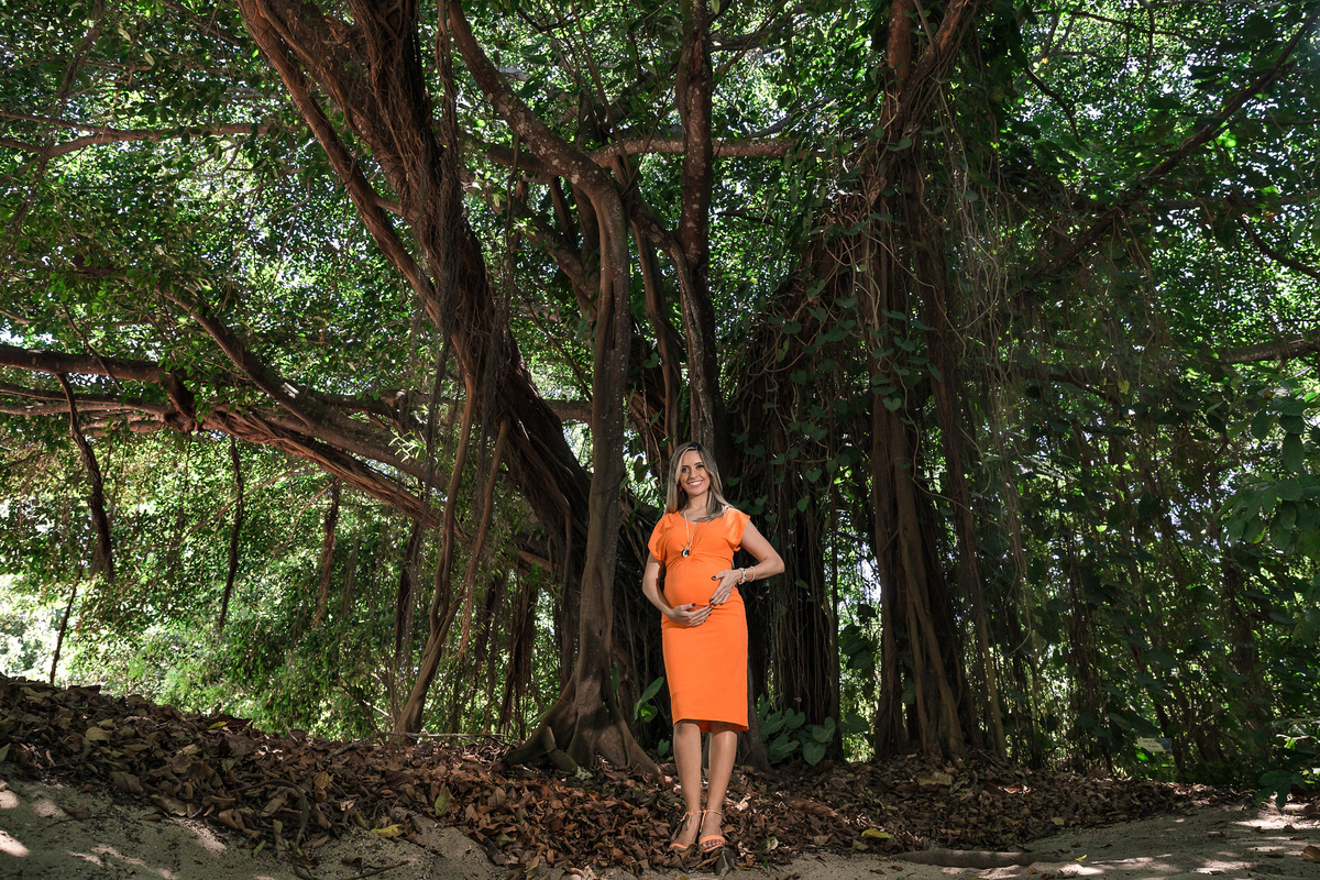 foto de gestante de vestido laranka e com as maos na barriga em frente a grande árvo no parque chico mendes no rio de janeiro em seu ensaio fotografico