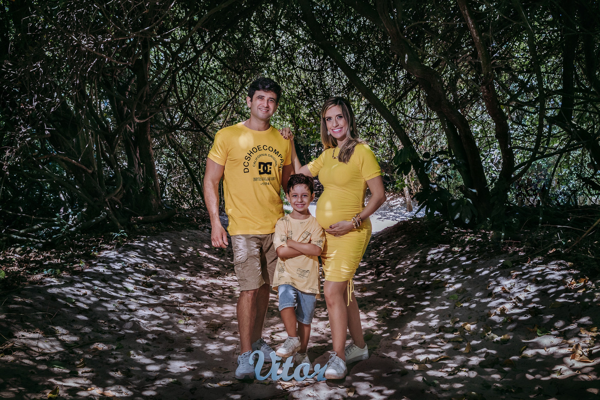 fotos de familia pai, mae com mao esquerda na barriga e direita sobre o ombro do marido e filho entre os dois e com o pé direito apoiado em placa com nome do irmao vitos em trilha do parque chico mendes no rj, todos de amarelo