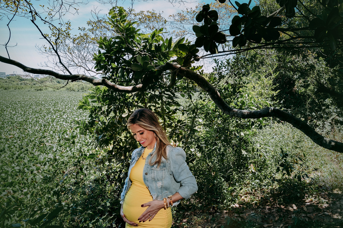 local com vegetação e um galho de árvore retorcido passando por tras de mulher gestante olhando pra barriga e com as maos na barriga e vestido amarelo e jaqueta