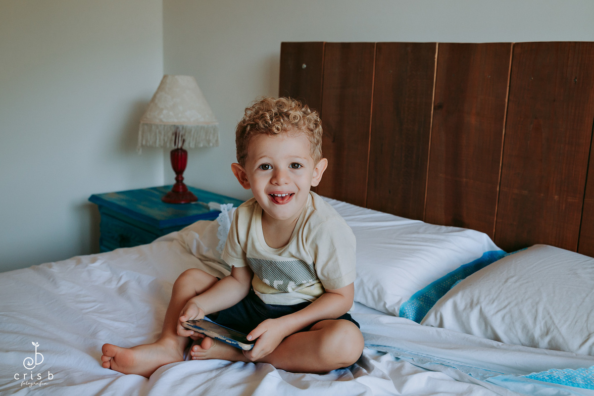 menino de 3 anos sentada na cama de casal com um celular na mão e dando careta