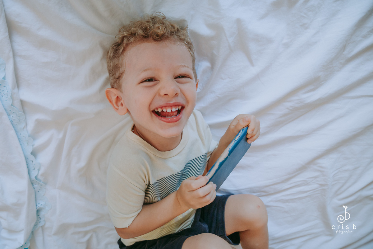 menino de tres anos deitado com um celular na mão e rindo pra camera