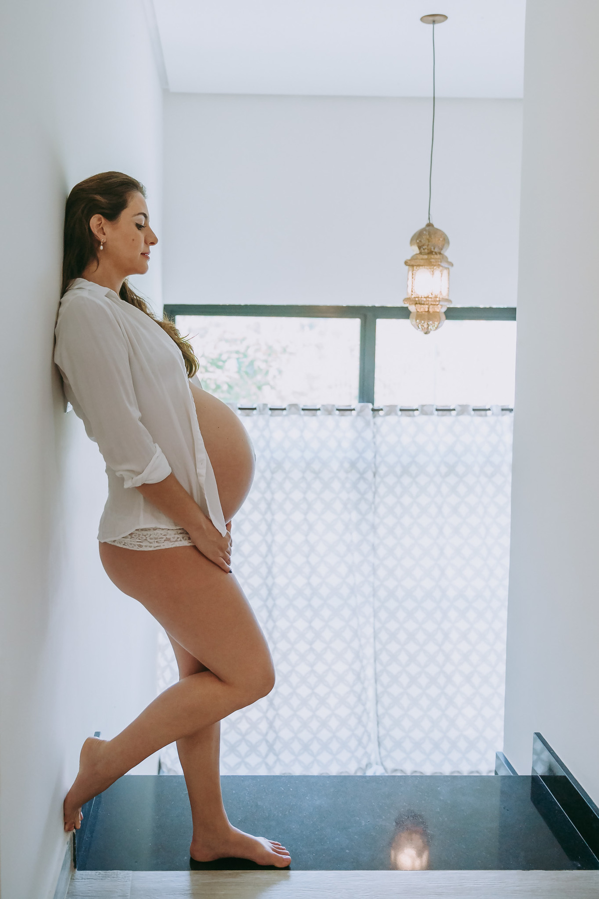 foto na vertical de gestante de corpo inteiro ela veste blusao branco e calcinha ela não olha pra camera está com semblante serio e olhando pra frente ao fundo janela e lustre