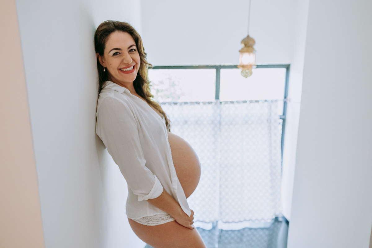 foto de ensaio de gestante com a gravida só de blusão branco no alto de uma escada e encostada numa parede e olhando sorrindo pra câmera ao fundo um janelão de vidro com cortina branca na frente