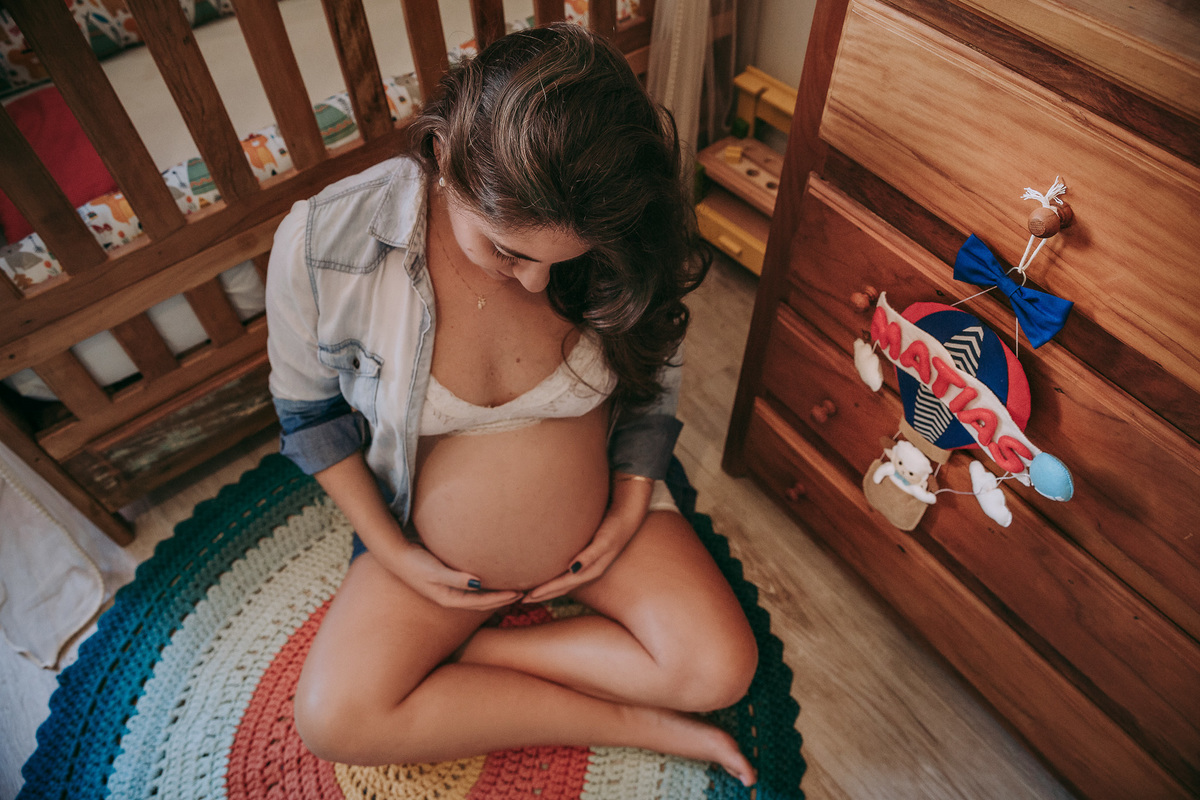 foto em plongee de gestante de sutiã e blusao sentada no chão do quarto do bebe  olhando para a barriga atyras dela o berço e ao lado esquerdo a comoda com um enfeite em forma de balão com o nome da criança