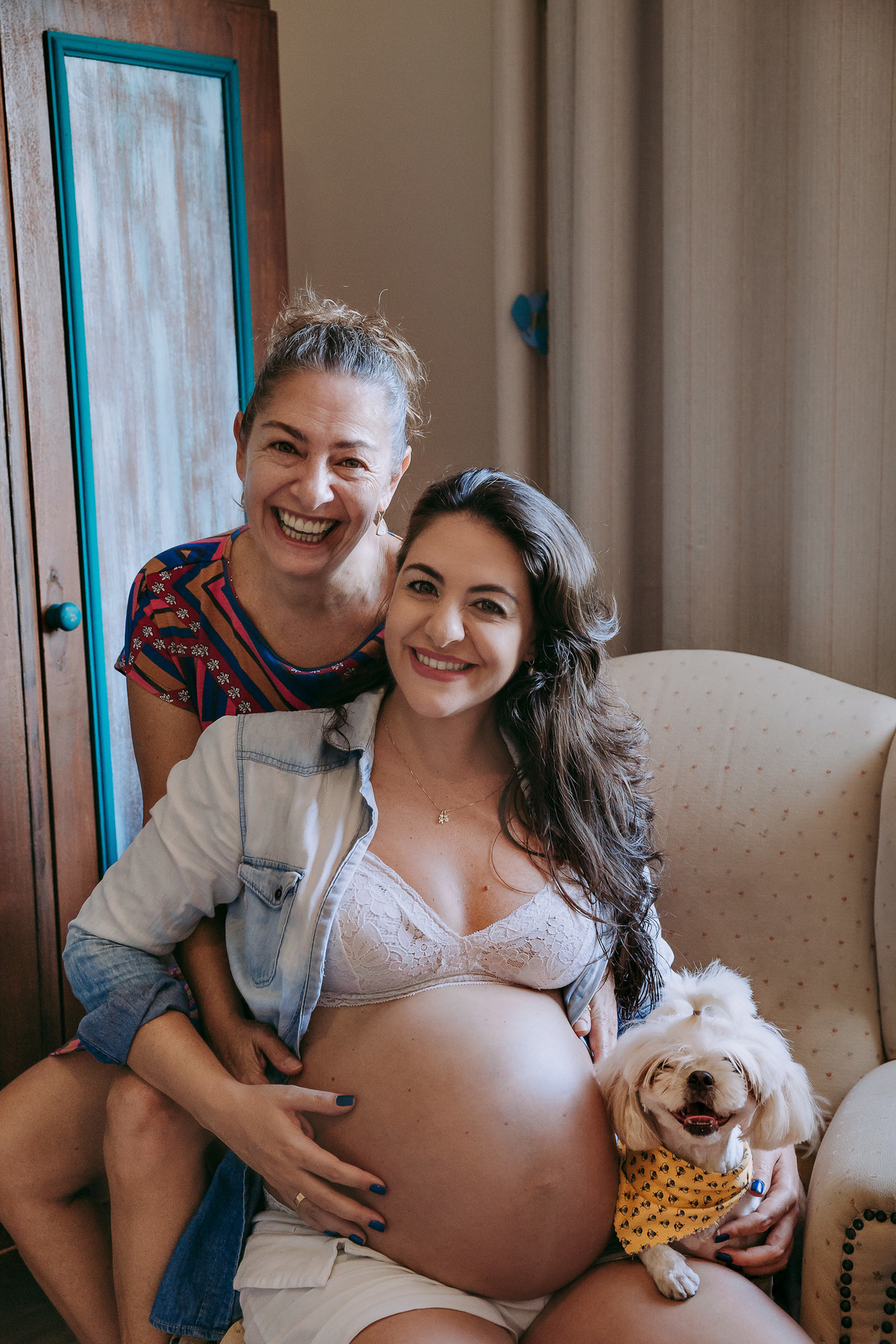 foto de gestante sentada no sofá acompanhada por sua mãe sentada no braço da poltrona e sua cachorrinha sentada em seu colo, todos sorriem pra foto
