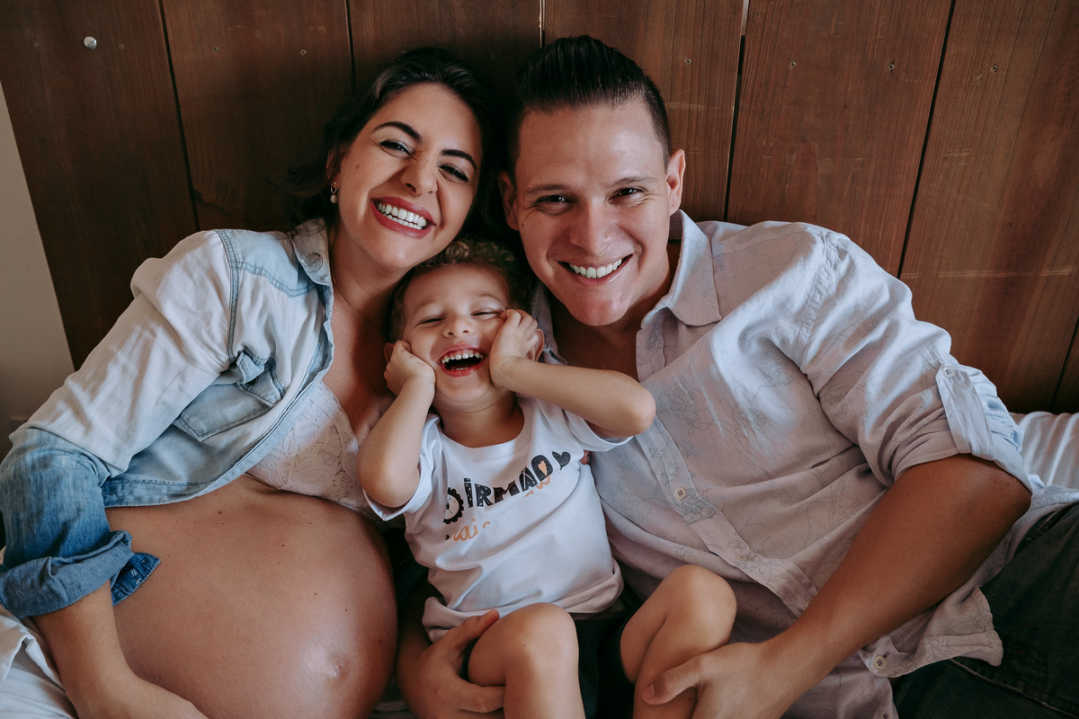 foto de familia, tres pessoas encostadas na cabeceira de uma cama de casal, foto em plongée, a gravida segura a barriga abraçada ao filho e marido, a criança, um menino de 2 anos, está sorrindo com as  maos na bochecha e o marido apoia sua mão esquerda n