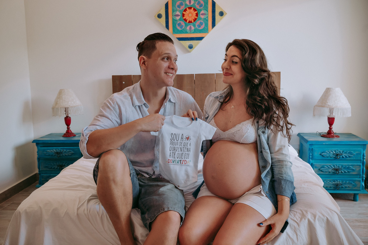 foto de casal sentados sobre a cama de seu quarto, ambos sorrindo, olhando um para o outro e segurando um body de bebê onde lê-se que a quarentena foi divertida, a mulher está gestante