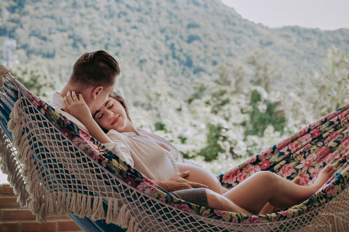 casal deitados na rede da varanda de sua casa num momento de relaxamento, marido beija a testa da gestante e ela de olhos fechados dá um leve sorriso
