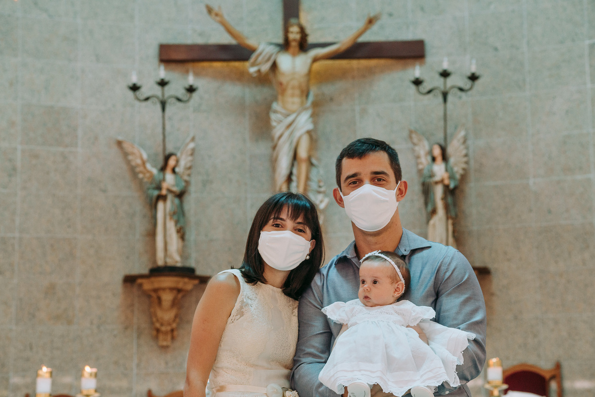 foto vertical de pai e mae com mascaras  em frente ao altar no batismo de sua filha pequena na Paróquia São Marcelino de Champagnat