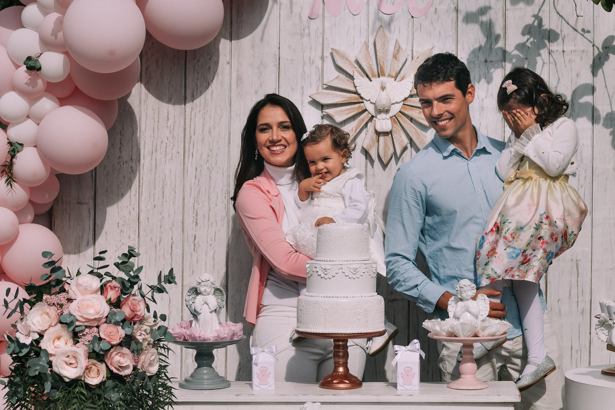 pai, mae e suas duas filhas posam para foto atras de mesa do bolo de batizado e a filha maior no colo esconde o rosto