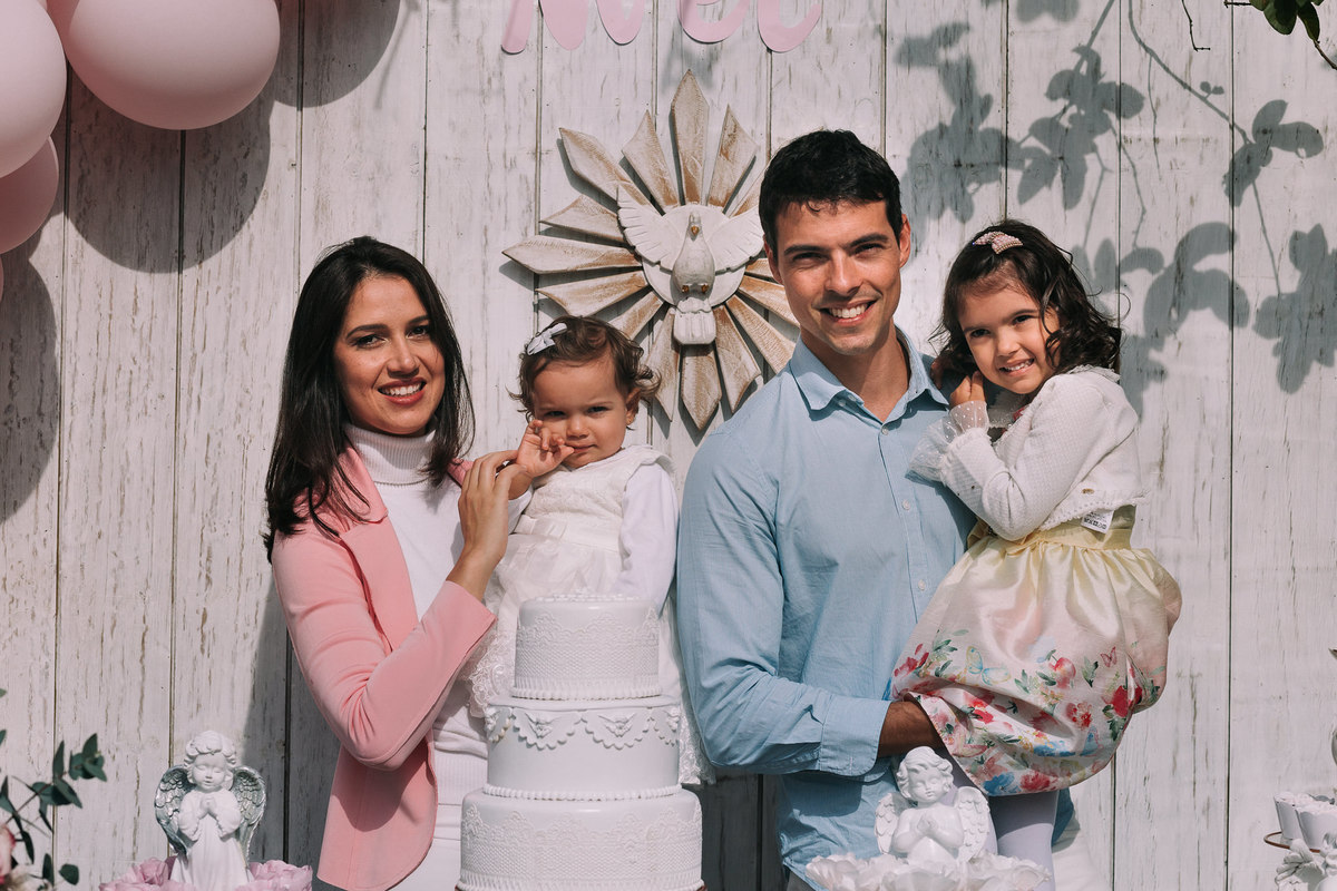 pai, mae e duas filhas posam para foto atras de mesa do bolo de batizado 