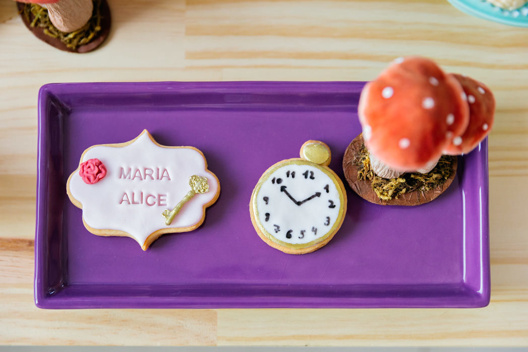 foto em plongée de uma bandeja roxa com um cogumelo, um biscoito escrito maria alice e um relogio, tudo no tema alice no pais das maravilhas