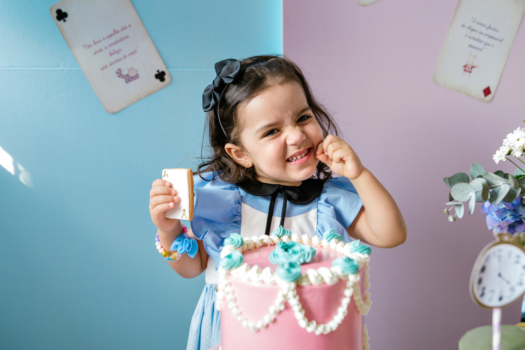 menina de tres anos atras de um bolo rosa. atras dela uma painel metade azul metade rosa com cartas de baralho coladas e escrito trechos da historia alice no pais das maravilhas