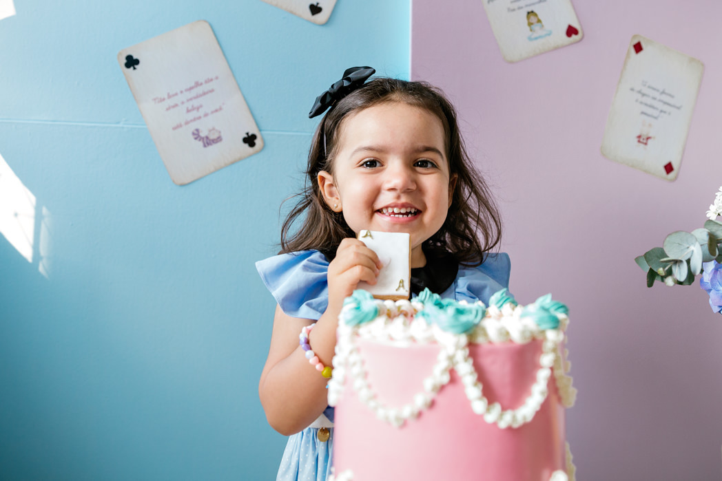 menina de vestido azul atras de seu bolo rosa com um painel atras dela nas cores rosae azul e com cartas de baralho impressas com trechos de alice no pais das maravilhas