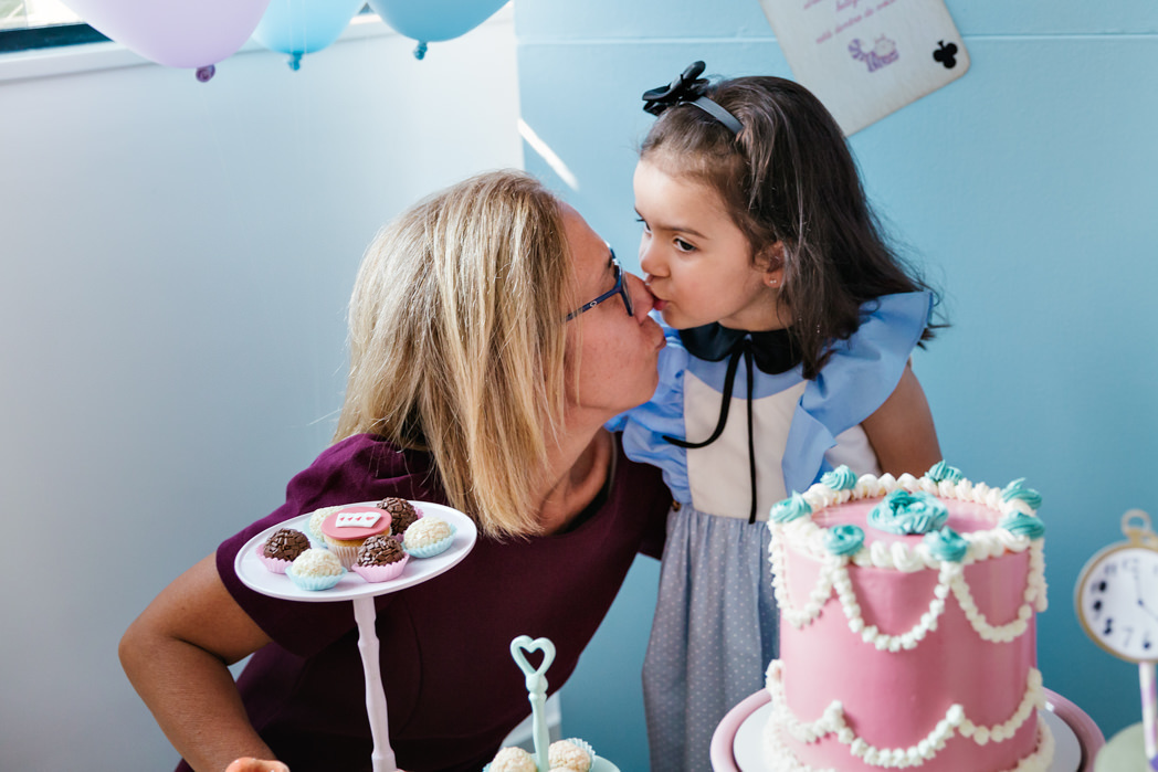 neta beija nariz de avó atras da mesa de bolo de seu aniversario com tema alice no pais das maravilhas