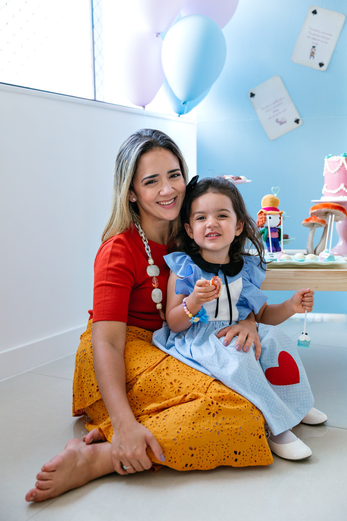 fotografia de mae e filha senstadas no chao em frente a mesa do bolo com tema alice no pais das maravilhas