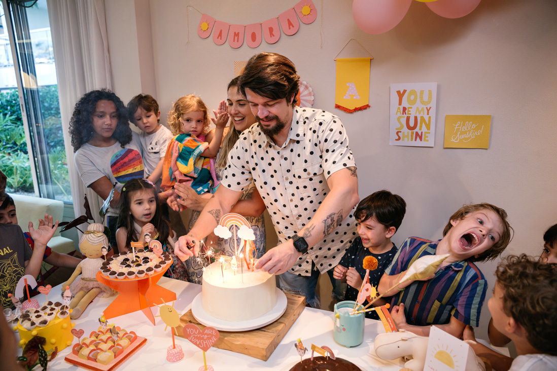 pai acebde as velas do bolo da festa da filha de 3 anos enqaunto o outro filho faz gracinhas