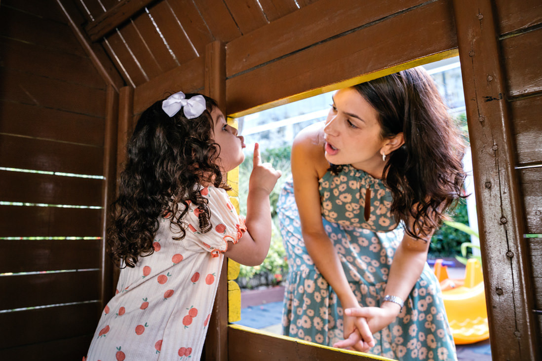 criança de tres anos dentro de casinha de parquinho pede silencio a mae com dedo indicador em punho