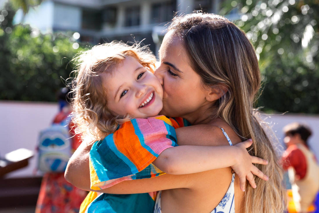 foto em contra luz de mae beijando forte a filha que sorri num dia de sol