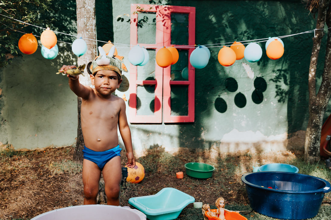 ao fundo um varal de baloes de gas nas cores laranja e azul, em primeiro plano menino mostra dinossauro
