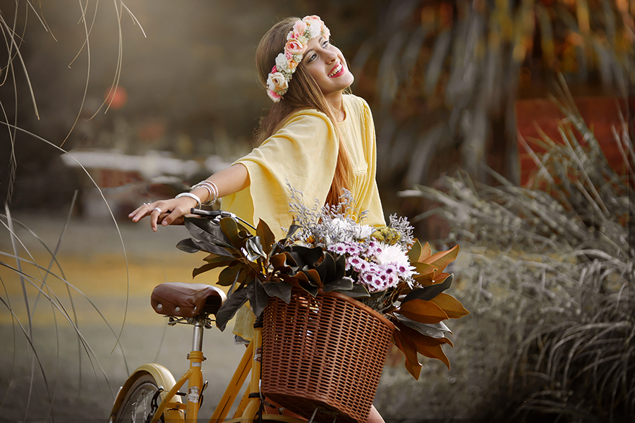 Quinceañera sonriente, mirando el cielo, con su bicicleta amarilla, su ramo de flores y su corona primaveral, disfrutando de un hermoso día de verano en un parque. Sesión quince años. Fotografía Barby Massei.