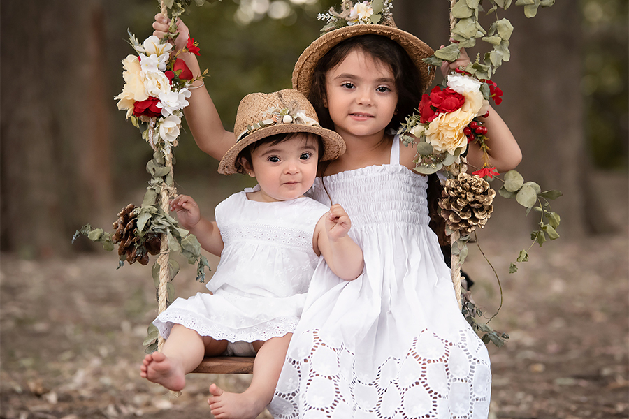 Hermanas con vestido blanco y sombrero de paja sobre hamaca con flores. Sesión de fotos para el book de Navidad en Santa Fe. Barby Massei fotografía