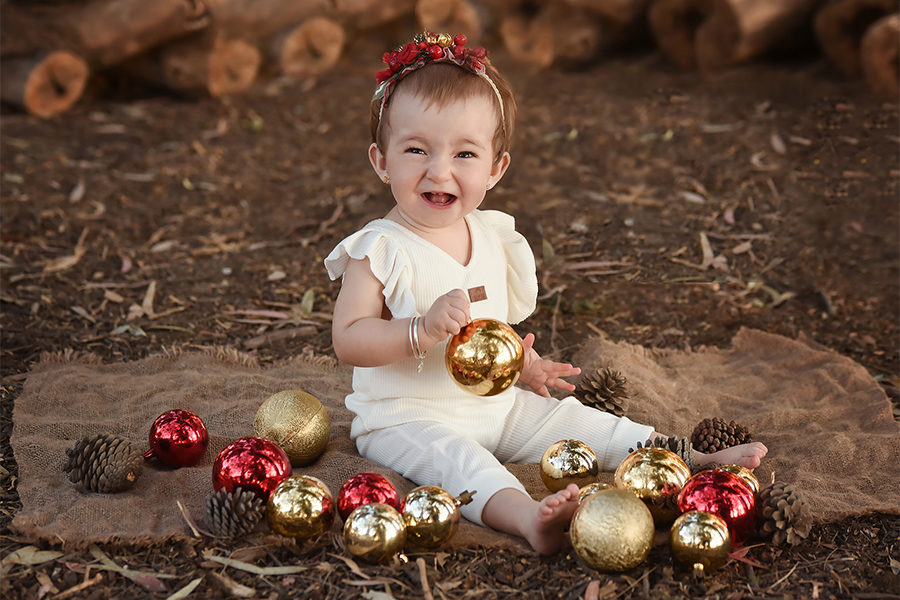 Niña con bolas de navidad rojo y dorado. Sesión de fotos navideñas en el campo. Vestido blanco y corona de flores. Barby Massei fotógrafa