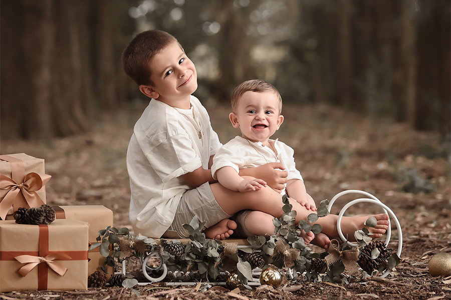 Hermanos en trineo navideño decorado con paquetes de regalo. Sesión de fotografía para el book de Navidad al atardecer en la naturaleza. Barby Massei foto Santa Fe
