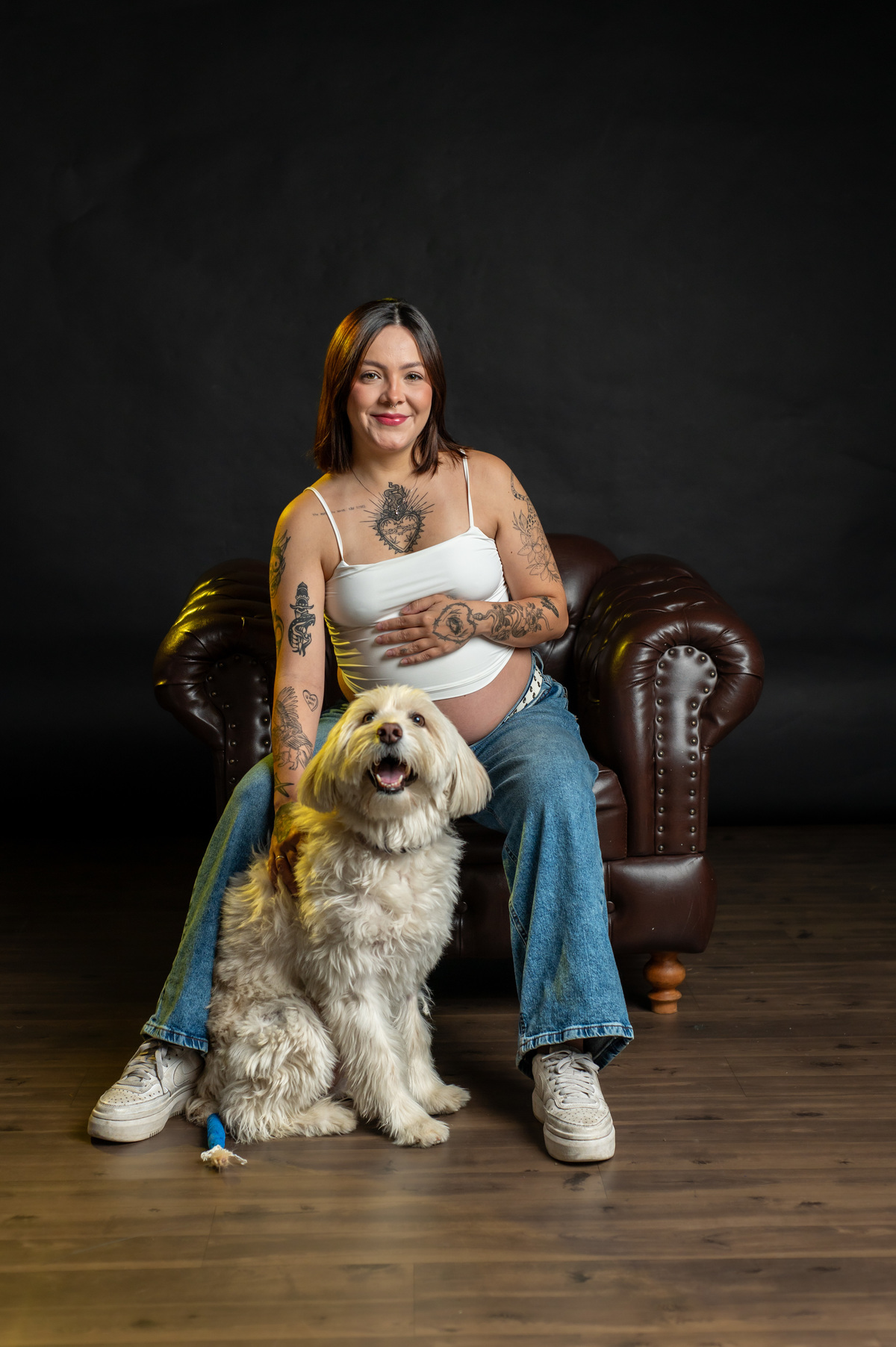 Gestante sentada em estúdio com cachorro, expressão leve e sorriso natural, ensaio de maternidade com pet, fotografia afetiva com luz suave e fundo escuro.