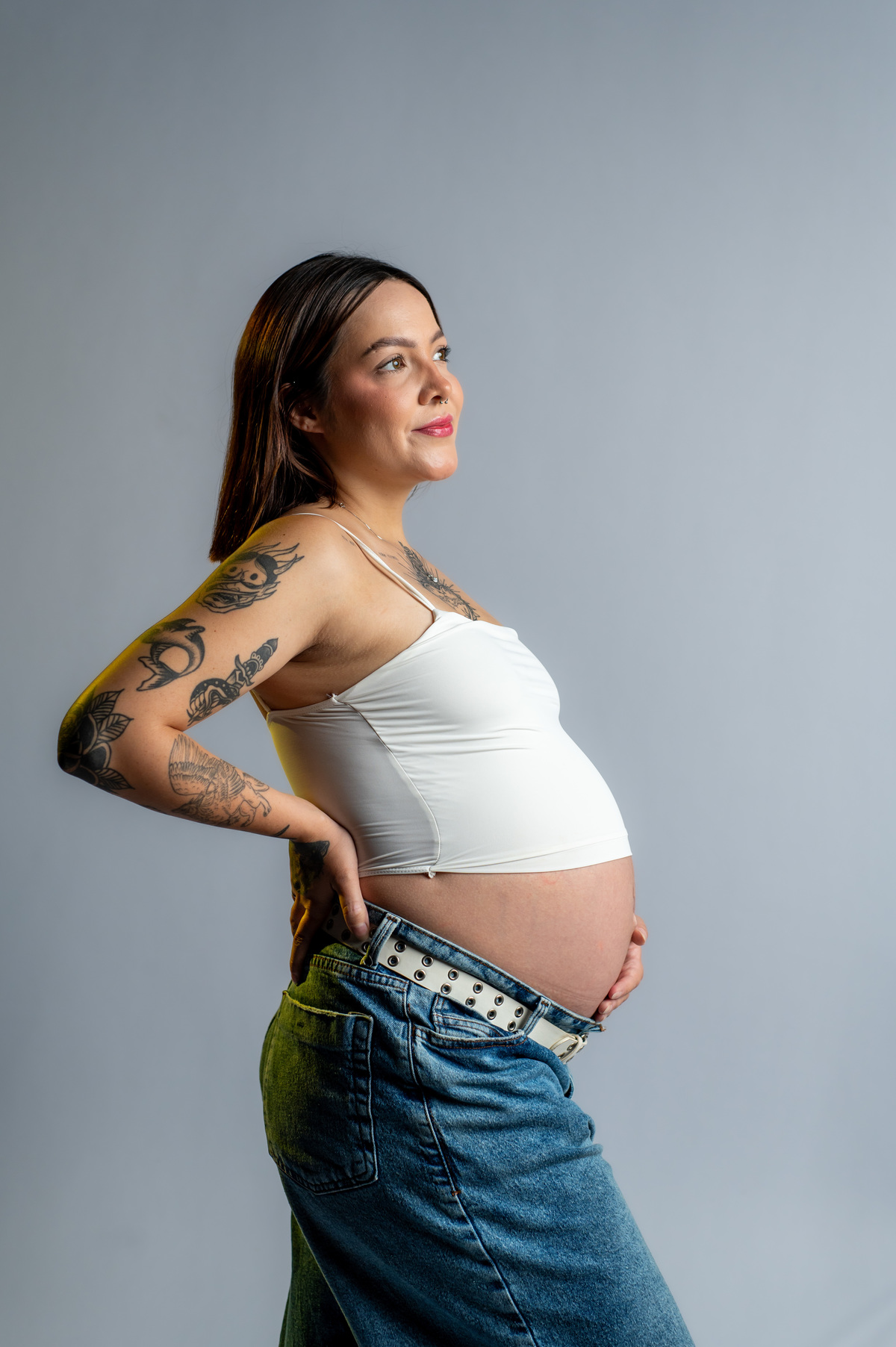 Retrato lateral de gestante em estúdio com fundo claro, destaque para a barriga, ensaio minimalista com luz suave e composição limpa.