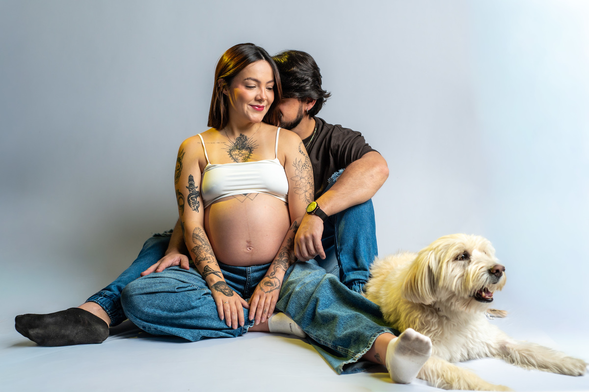 Casal gestante sentado no chão de estúdio com cachorro ao lado, composição leve e natural, ensaio com interação familiar e clima afetivo.